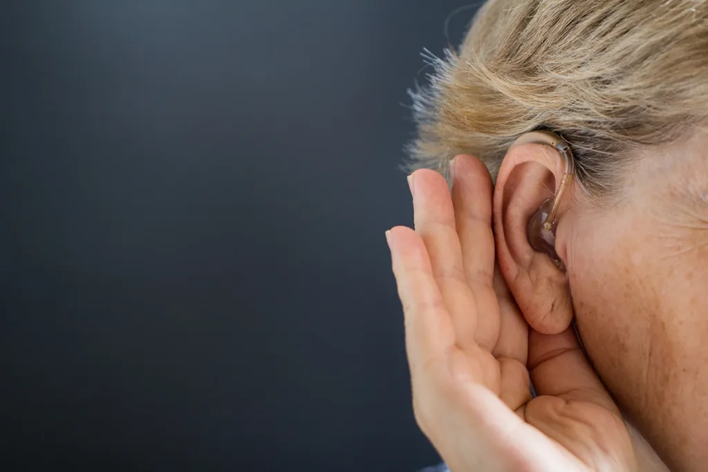 Close up of a middle aged person wearing a behind the ear hearing aid, cupping their hand around their ear to hear better, suggesting the need to replace hearing aids 