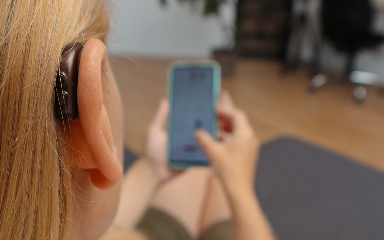 A close up of a woman wearing a behind-the-ear (BTE) hearing aid and looking down at a hearing aid app on her smartphone.